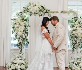 Bride in white lace and groom in beige suit hold hands under a floral arch at an elegant New Orleans wedding.
