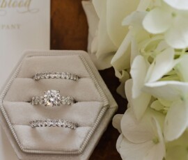 Three diamond rings on a hexagonal white velvet box beside white hydrangeas and gold script paper—stylish Louisiana wedding inspo.