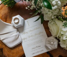 Wedding invitation with gold and black lettering, surrounded by white heel, rings, and bouquet on wood surface.