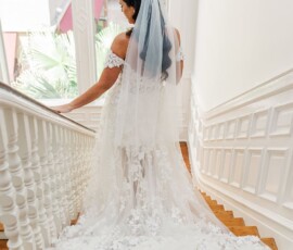 Bride in lace dress and veil on wooden stairs, looking back toward a large window, evoking romantic New Orleans wedding style.