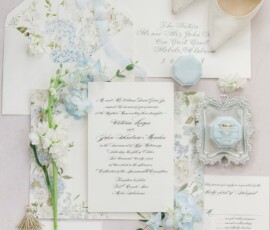 Flat lay of floral wedding invitations, pearl heels, flowers, blue ring box, gold jewelry, and envelope on a pale surface.