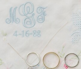 Three rings on white embroidered cloth with blue “MGF” initials, 4-16-88 date, and pastel floral designs—timeless wedding detail.