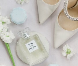 Elegant flat lay: jeweled white heels, Chanel No. 5, blue ring box with rings, and white flowers on a light surface.