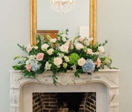 Elegant white marble fireplace with gold mirror, floral arrangement, and crystal chandelier reflected above—New Orleans wedding decor.