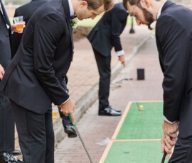 Four men in suits play outdoor mini-golf on a green mat, with trees, a street, and wedding reception vibes in the background.