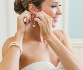 Bride in strapless white dress smiles, puts on dangling earring, pearl bracelet and neat bun at New Orleans wedding.