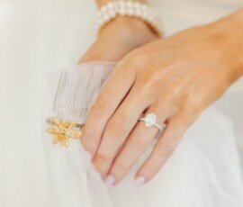 Woman’s hand with pearl bracelet and diamond ring holds white tulle with gold floral brooch—elegant Louisiana wedding inspo.