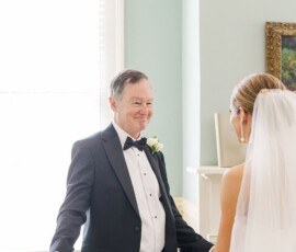 A groom in a black tuxedo smiles with open arms at a bride in white, sharing a joyful moment in a bright wedding room.