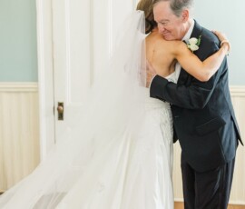 Bride in white gown hugs older man in black suit, both smiling indoors by a white door at their Louisiana wedding.
