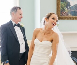Bride in strapless white dress laughs beside older man in tuxedo, joyful Gulf South wedding scene with painting in bright room.