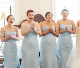 Six bridesmaids in light blue dresses smile and clap at a bride in white in a bright, sunlit Louisiana wedding room.
