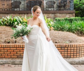 Bride in strapless white gown and veil holds bouquet on brick path by historic Louisiana building with large windows.