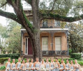 Sixteen women in matching light blue dresses pose with bouquets by a tree and historic brick house at a Louisiana wedding.