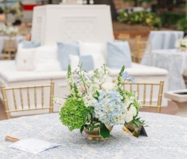 Round table with blue and white floral cloth, blue-green-white flower centerpiece, gold chairs, white sofas, lush greenery.