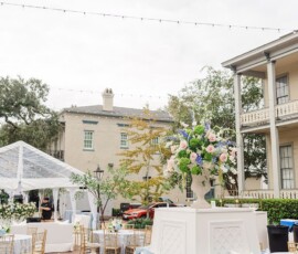 Outdoor wedding venue with blue-clothed tables, gold chairs, white benches, floral centerpiece, and string lights between historic buildings.