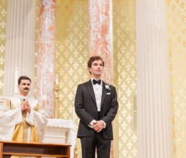 Groom in black tuxedo stands at grand church altar with ornate columns; priest in gold robes behind him, New Orleans wedding.