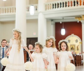 Flower girls in white dresses and floral crowns walk hand-in-hand with bouquets down a marble aisle in an ornate Louisiana church.