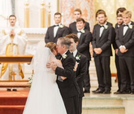 Bride in white gown hugs older man at church altar; groomsmen and priest stand behind at heartfelt Louisiana wedding.