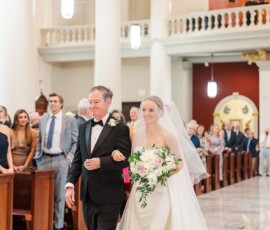 Bride in white gown and veil smiles, walking down church aisle with older man as guests watch elegant wedding ceremony.