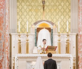 Bride and groom kneel at a church altar as a priest reads beneath a crucifix during their wedding ceremony.