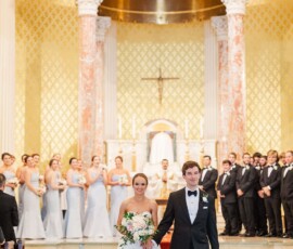 Bride and groom smiling as they walk down the aisle in a grand Louisiana church, bridal party in light attire behind them.
