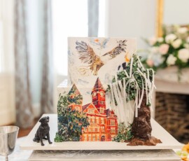 New Orleans wedding cake with painted building, eagle, trees, black dog figurine, white streamers on silver stand.