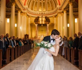 Bride and groom kiss in a grand church aisle, surrounded by guests. She wears white with a veil; he’s in a tuxedo.
