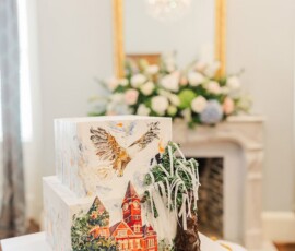 Two-tiered cake with castle, owl, tree, and black cat on ornate stand amid flowers and elegant fireplace—romantic wedding inspo.