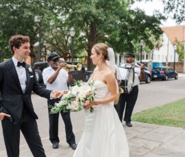 Bride and groom smile outdoors at their Louisiana wedding as musicians play trumpet and drums; trees and cars in background.
