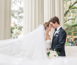 Bride and groom touch foreheads outside, her veil flowing by classic white columns and lush New Orleans greenery.