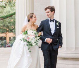 Bride in white gown and groom in black tuxedo smile and walk by white columns and greenery at a classic Gulf South wedding.