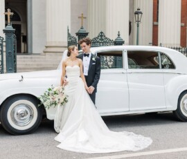 Bride and groom in wedding attire smile at each other by a white vintage car, outside elegant columns and iron gates.