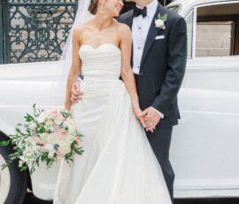Bride in white dress and groom in black tux kiss forehead, holding hands by a white car with her bouquet at New Orleans wedding.