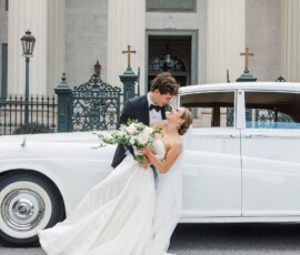 Bride and groom smile by a white vintage car at their New Orleans wedding, with columns and iron gates in the background.
