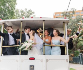 Bride, groom, and wedding party celebrate on a flower-adorned white trolley in New Orleans city street.
