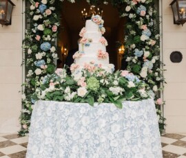 Tiered white wedding cake with pastel flowers on a lace table, surrounded by floral decor at an elegant New Orleans venue entrance.
