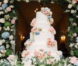 Four-tier white wedding cake with pink and blue flowers, lush greenery, and a floral arch at a romantic Louisiana wedding.