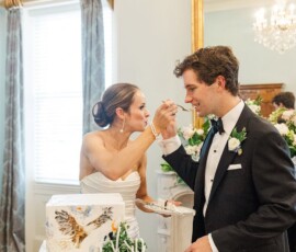 Bride in strapless dress feeds cake to groom by an owl-themed cake at a floral Louisiana wedding with chandelier.