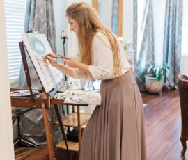 Woman in a white blouse and mauve skirt paints at an easel in a bright, elegant room with wooden floors and large windows.