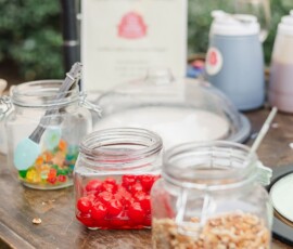 Three glass jars with walnuts, cherries, and gummy bears sit on a wooden table with sauces and greenery in the background.