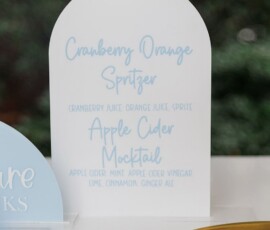 White drink sign displays Cranberry Orange Spritzer and Apple Cider Mocktail, with gold tray and greenery in the background.