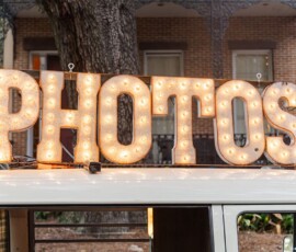 Light-up marquee letters spelling PHOTOS sit atop a white vehicle at a New Orleans wedding, with trees and a building behind.