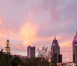 Downtown skyline at sunset with pink-orange clouds, pointed buildings, a lit tower, and trees—Gulf South wedding inspiration.