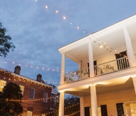 String lights glow between two elegant houses at dusk, creating a warm, inviting Louisiana wedding atmosphere.