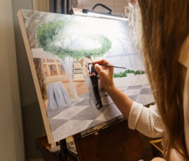 An artist with long hair paints a Louisiana wedding scene of a couple dancing under trees, with paint and brushes nearby.