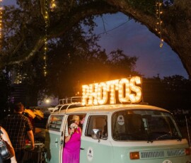 Mint-green vintage VW photo booth van under string lights at a New Orleans wedding, with guests nearby and a woman in pink inside.