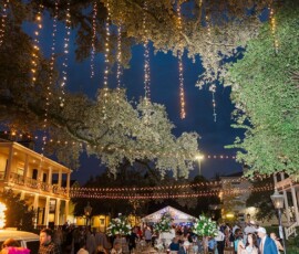 Lively Louisiana evening wedding outdoors, string lights, floral centerpieces, guests at tables, historic buildings behind.