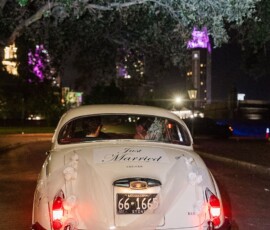 Vintage white car with “Just Married” sign, streamers, and pom-poms drives away at night under city lights in New Orleans.