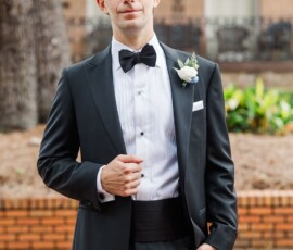 Man in black tuxedo adjusts jacket outdoors, with greenery and brick building behind—New Orleans wedding ready.
