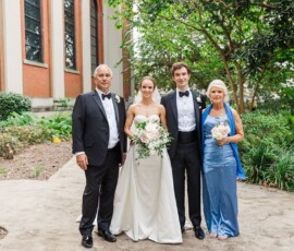 Bride, groom, and parents in formalwear smile outdoors by a brick building—a charming Gulf South wedding moment.
